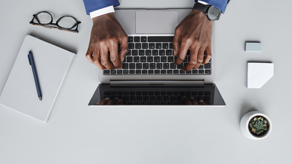 Business professional typing on a laptop at a clean desk with notebook, pen, glasses, plant, and modern office accessories.