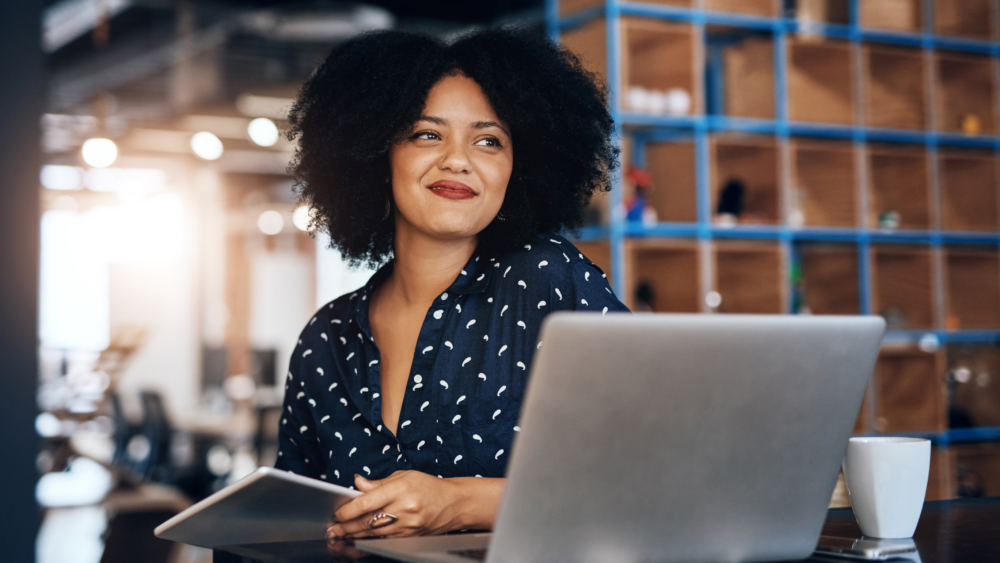 Smiling businesswoman working on a laptop in a modern office, holding a tablet and enjoying coffee.