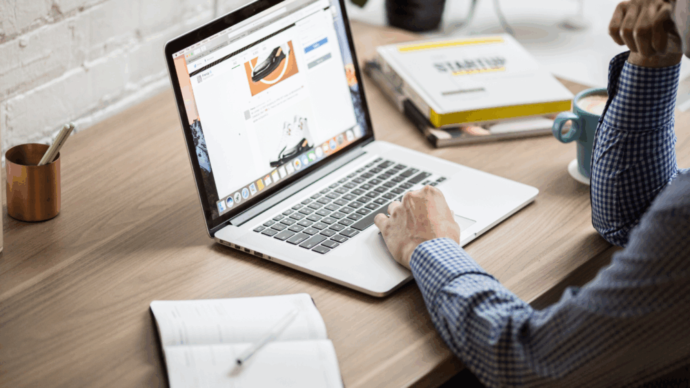 Professional working on a laptop at a wooden desk with notebook, coffee mug, and business documents.